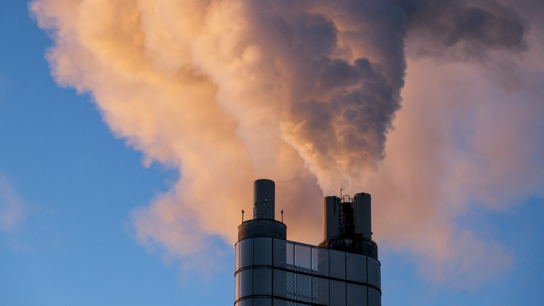 Røyk velter opp fra en industriell skorstein mot en blå himmel og skaper en stor forurensningssky.