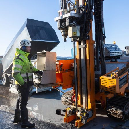 En arbeider betjener tungt boremaskineri på en travel byggeplass, kledd i vernehjelm og synlig jakke. En lastebil ruver i bakgrunnen, omgitt av utstyr, ettersom innsatsen for å minimere utslipp er tydelig i hele prosjektområdet.