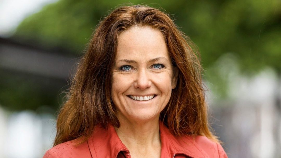 En person med langt brunt hår som smiler, iført en livlig rød skjorte, står midt i en utendørs setting. Det uskarpe grøntområdet i bakgrunnen antyder subtilt naturens rolle i å bekjempe klimagassutslipp.