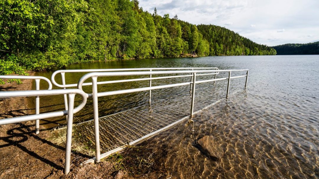 Metallrekkverk leder inn i en klar innsjø, en av regionens fineste badeplasser, omgitt av tett grønn skog under overskyet himmel.