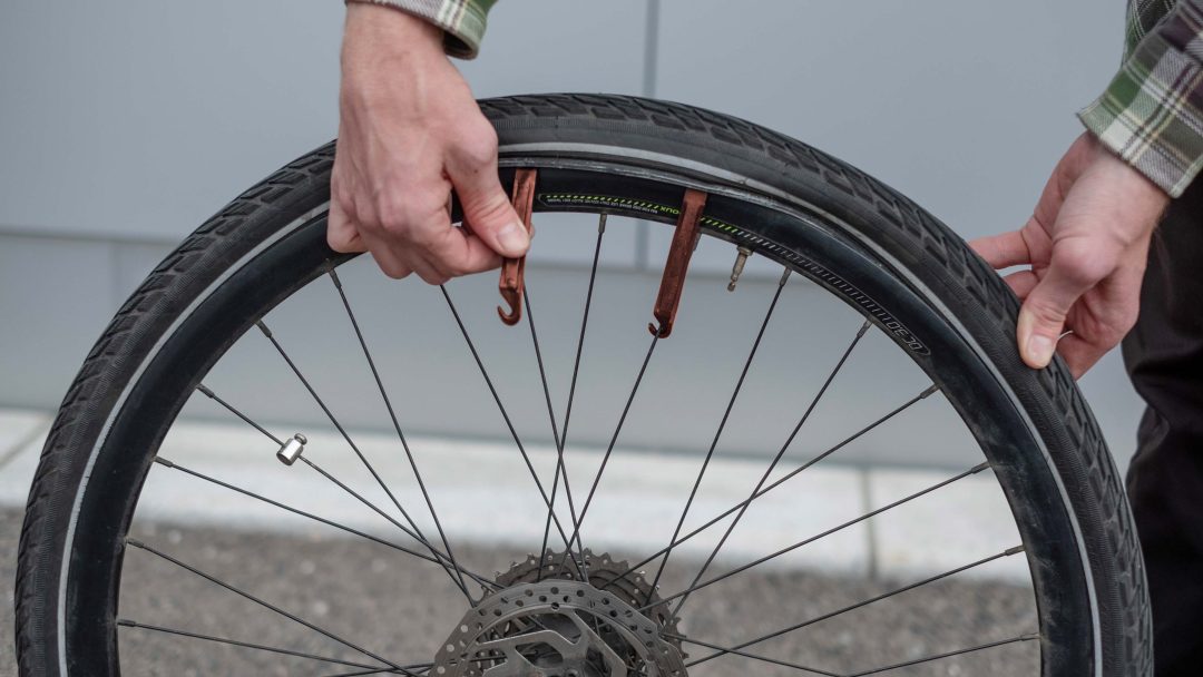 En person demonstrerer hvordan man skifter dekk ved å fjerne et sykkeldekk med brune dekkspaker på utendørs fortau.