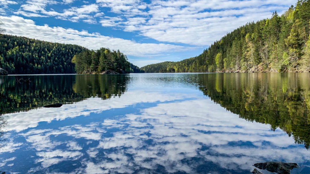 En rolig innsjø med skogkledde åser som speiler seg i vannet, under en himmel med spredte skyer, tilbyr perfekte badeplasser for de som søker ro.