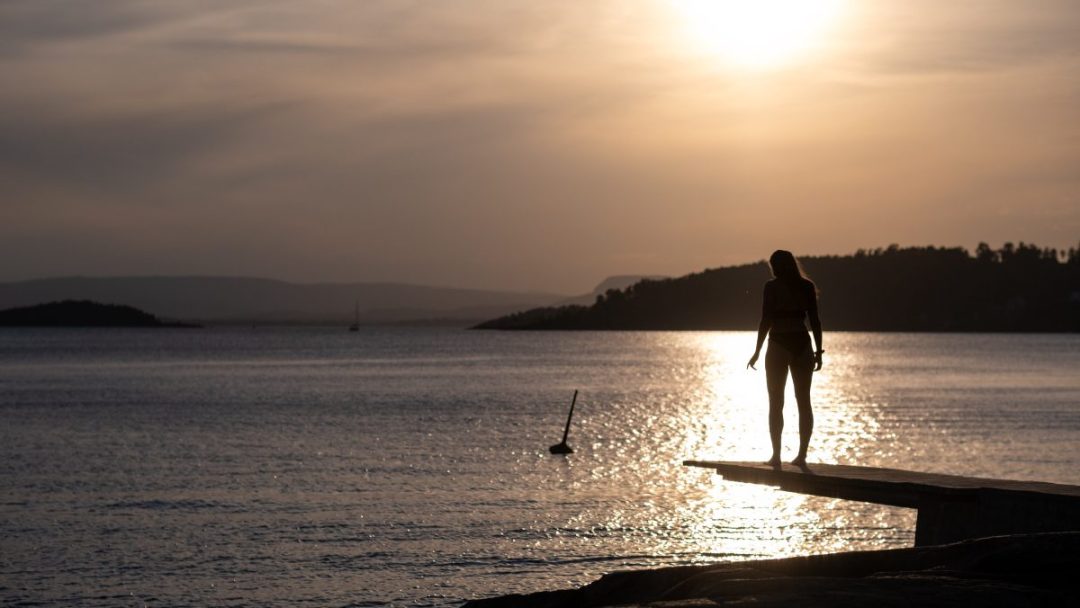 Silhuett av en person som står på en brygge ved en av de rolige badeplassene og stirrer på solnedgangen over et stille hav med fjerne åser.