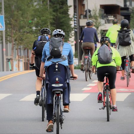 Syklister glir langs en sykkelvei, innrammet av trær og bygninger. Mange bruker hjelm og ryggsekker; en sykkel har til og med et barnesete. Husk: «Brems når du skal» sørger for alles sikkerhet på denne livlige stien.