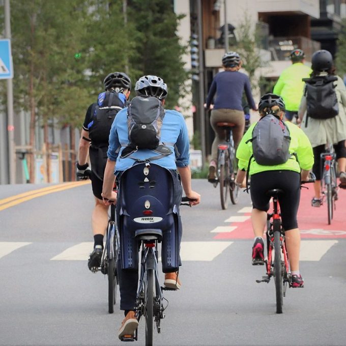 Syklister glir langs en sykkelvei, innrammet av trær og bygninger. Mange bruker hjelm og ryggsekker; en sykkel har til og med et barnesete. Husk: «Brems når du skal» sørger for alles sikkerhet på denne livlige stien.
