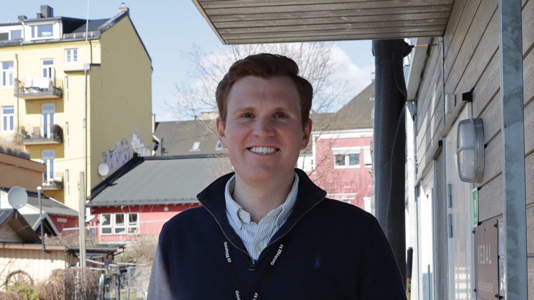 En person som smiler utendørs, står foran en trebygning. Bakgrunnen viser en gul og rød bygning med noen trær og klar himmel – en perfekt dag ubesmittet av klimagassutslipp.