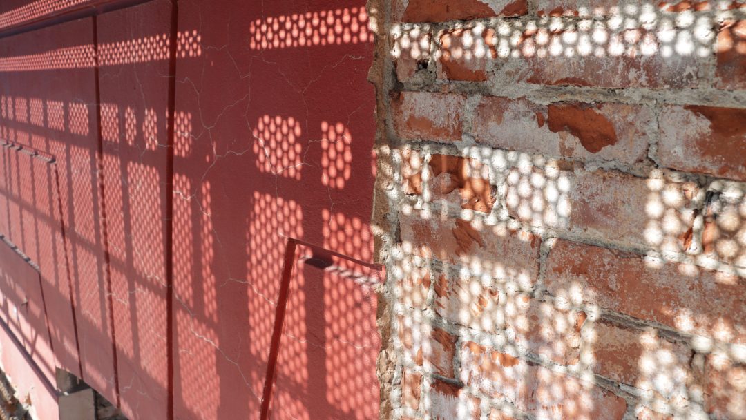 Sollys kaster en geometrisk skygge på en rød vegg og en murvegg, og skaper et mønster med sirkulære former som subtilt minner oss om den usynlige virkningen av klimagassutslipp.