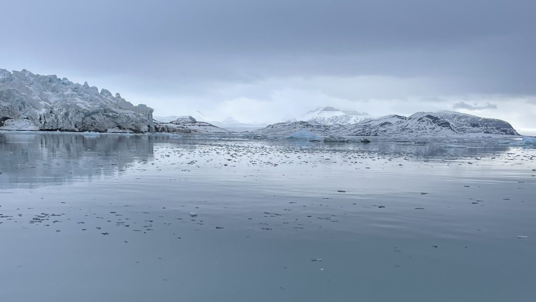 Et rolig islandskap med en isbre til venstre, en reflekterende vannmasse i forgrunnen og snødekte fjell i bakgrunnen under en overskyet himmel.