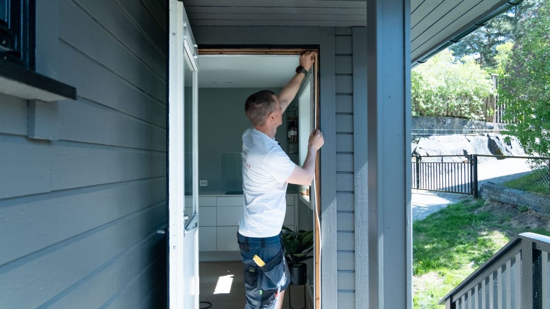 Person i hvit skjorte og blå bukser jobber på montere tetningslister for en dørkarm til et grått hus. Solbelyst utendørsscene med grøntområder og en sti i bakgrunnen, som fremhever den grundige innsatsen som er involvert.