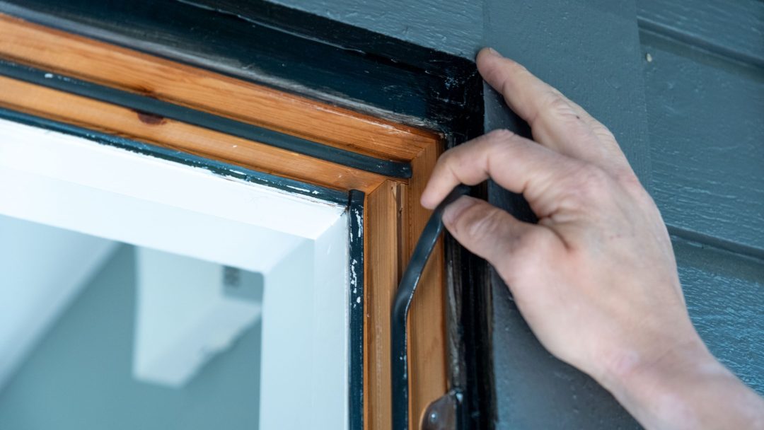 En hånd som ekspertjusterer svart tetningslister på et treinnrammet vindu med en malt kant.