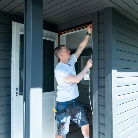 En person i hvit t-skjorte og shorts jobber dyktig med husets eksteriør og monterer tetningslister nær en dør. Sollys lyser vakkert opp scenen mens de forsiktig håndterer ledningen.