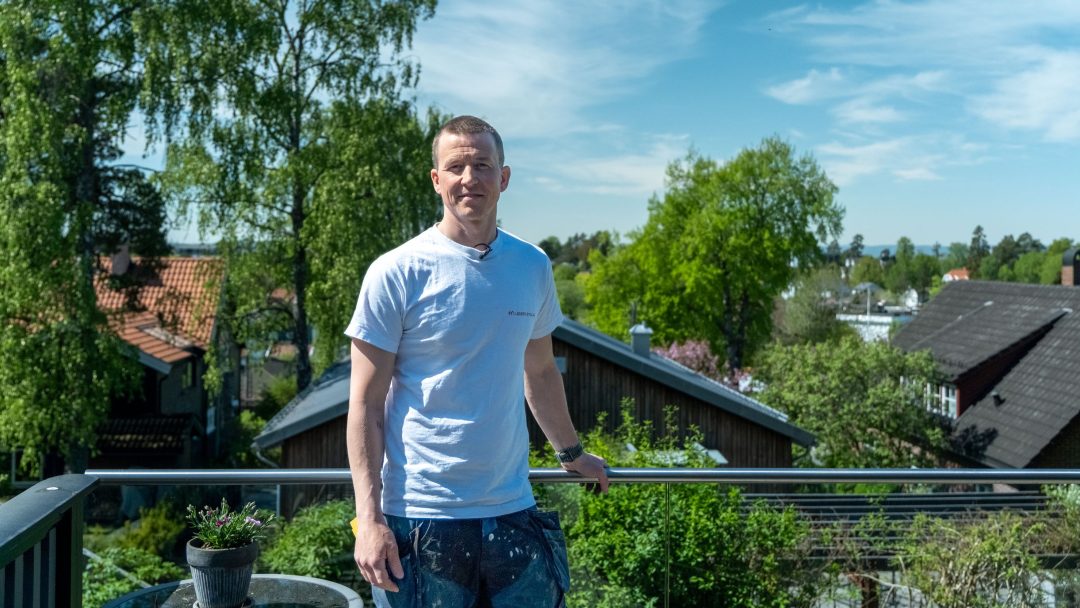 En mann i en hvit t-skjorte står på en balkong med en vakker utsikt over trær og hustak under klar himmel, og vurderer hvor han skal montere tetningslister for ekstra komfort.
