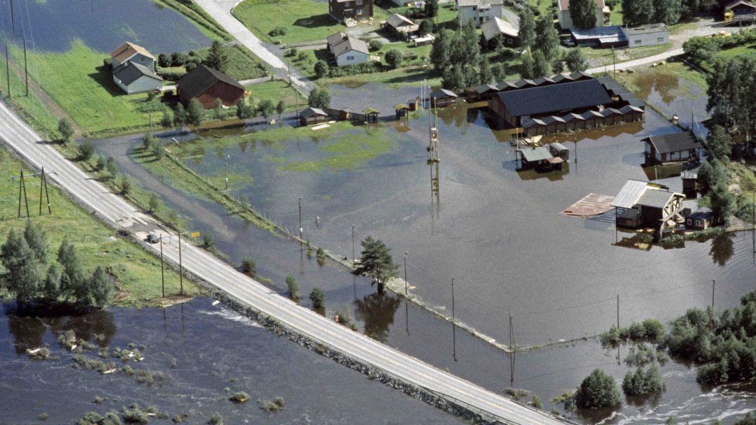 Luftfoto av et oversvømmet landlig område med nedsenkede hus, veier og vegetasjon. Vann dekker det meste av landskapet.