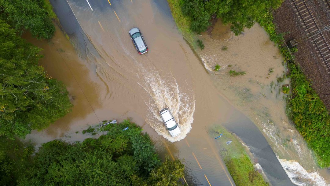 Luftfoto av to biler som kjører gjennom en oversvømt vei omgitt av trær, med vann som sprer seg til området i nærheten.