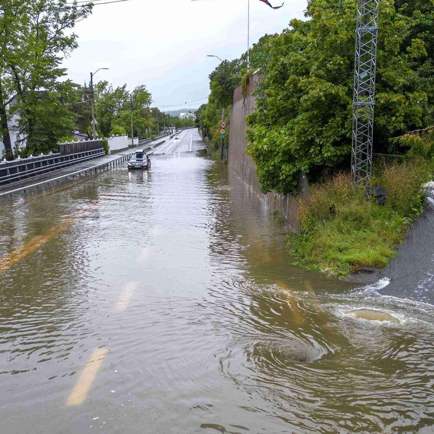 En bil navigerer på en oversvømt vei i Norge, omgitt av trær og bruksstolper. Vann fra nyere klimaendringer renner ned en skråning til høyre under en overskyet himmel.