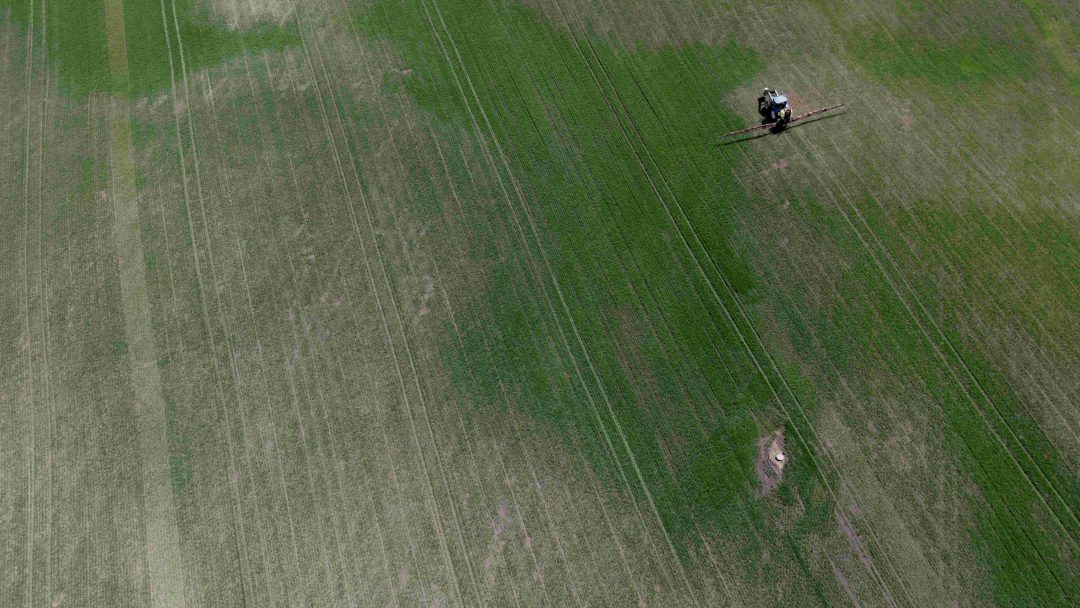 Luftfoto av et stort grønt jordbruksfelt med en traktor som sprøyter avlinger i øvre høyre del.