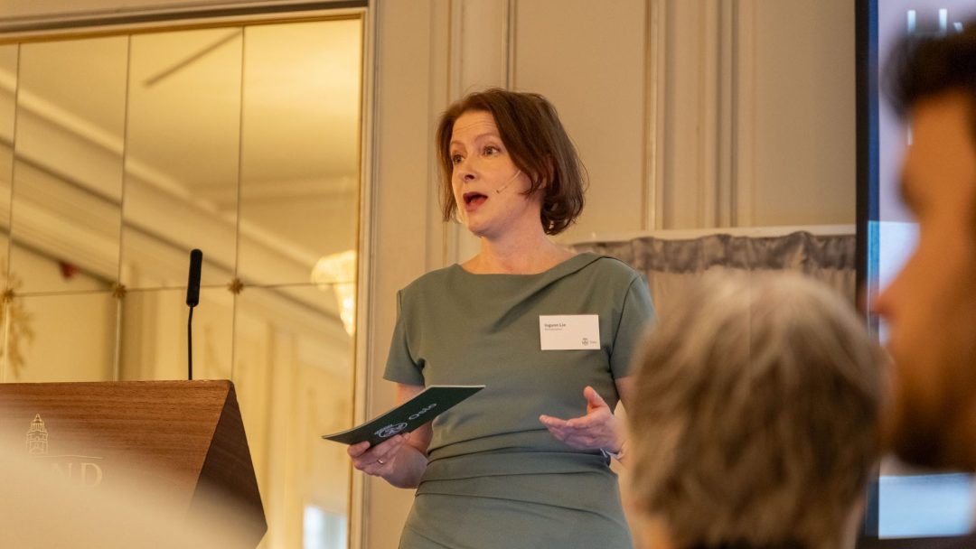 En kvinne i grønn kjole snakker engasjert ved et podium om klimaregnskap, mens hun holder et hefte i konferanserommet. Oppmerksomme publikummere er synlige i forgrunnen.