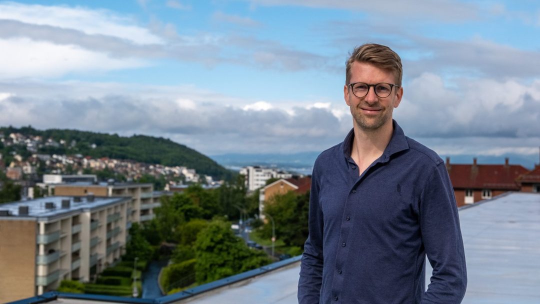 En mann i briller og blå skjorte står på et tak utsmykket med solceller, mot et bakteppe av bybilde og grøntområder under en delvis overskyet himmel.