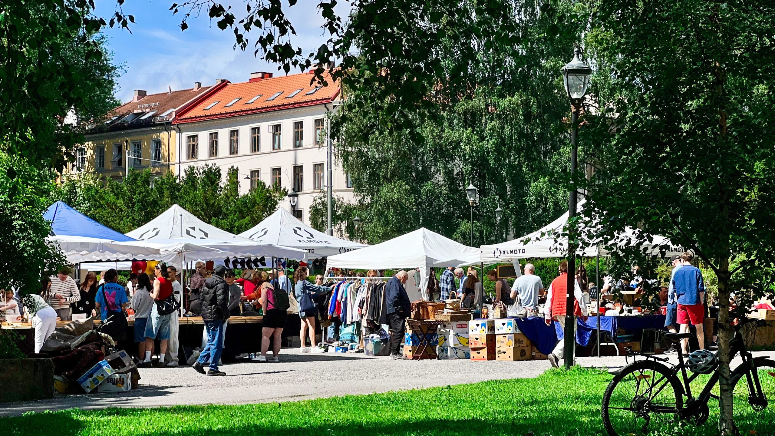 Folk som surfer på boder på et utendørsmarked i en park, på en skattejakt for unike funn. Teltene viser klær og diverse gjenstander, mens sykler og trær rammer inn den livlige scenen. Bygninger er synlige i bakgrunnen, noe som bidrar til atmosfæren av utforskning og oppdagelse.