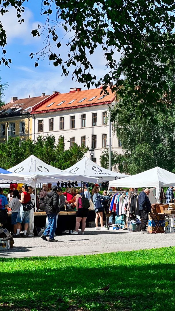 Shoppere nyter en skattejakt gjennom utendørsmarkedet, og utforsker boder under hvite telt fylt med unike skatter. I bakgrunnen titter sjarmerende bygninger og frodige trær under en delvis overskyet himmel.