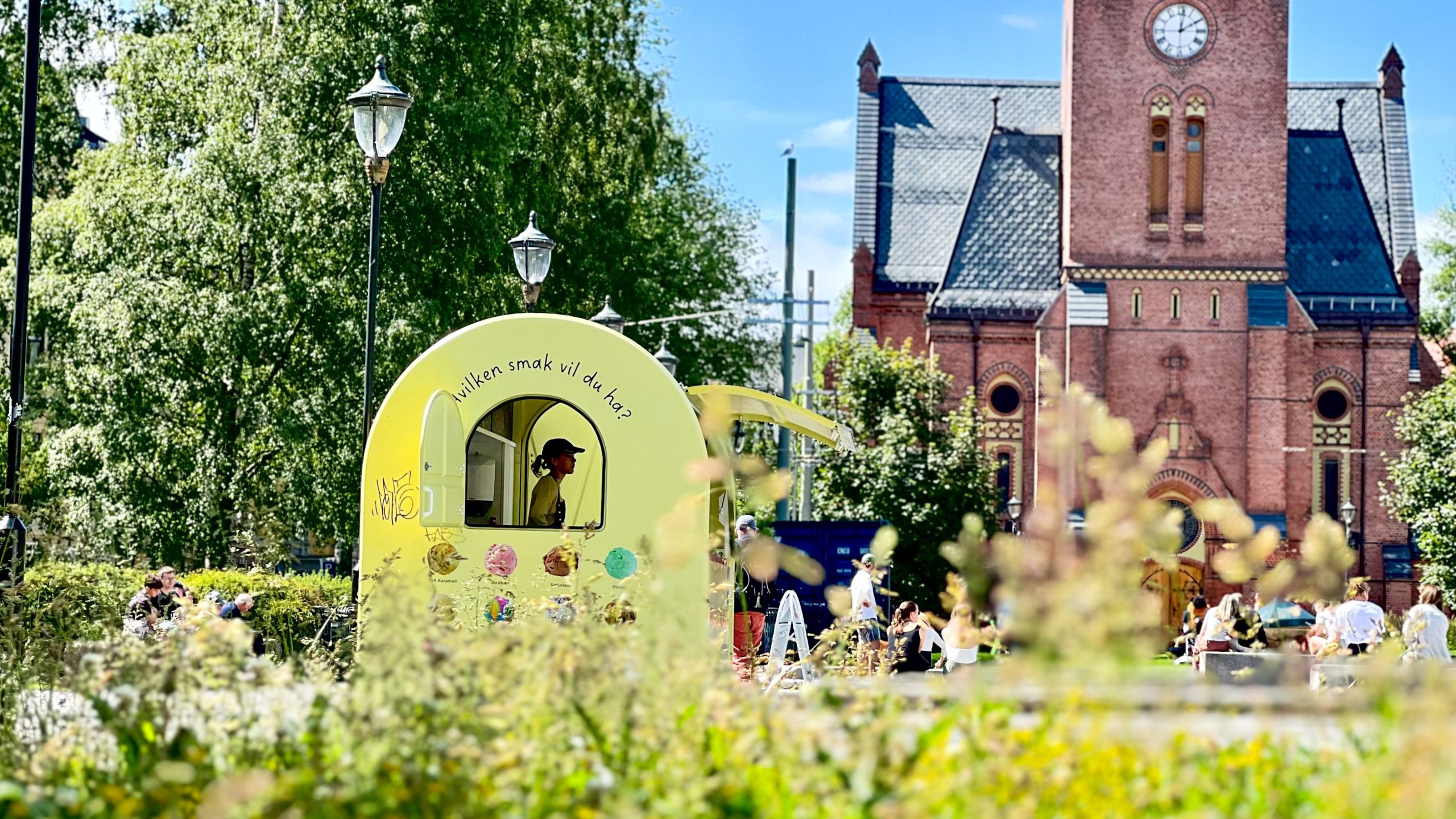 Et lite gult iskrembord med en person inni er satt opp i parken mens skattejaktdeltakerne samles i nærheten for å søke sin neste ledetråd. En murbygning med et klokketårn fungerer som et ikonisk bakteppe for dette morsomme eventyret.