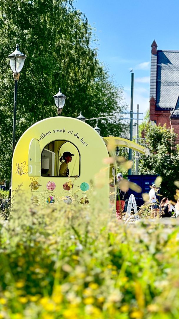 En person inne i en lysegrønn foodtruck med en Vilken smak vil du ha? skilt, omgitt av frodige grøntområder og en bygning i bakgrunnen, legger ut på en kulinarisk skattejakt for å glede smaksløkene.