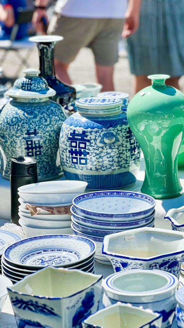 En samling av blå og hvite keramikkfat og vaser, sammen med en grønn vase, står kunstferdig ut på et bord utendørs, som minner om en skattejaktskatt som venter på å bli oppdaget.