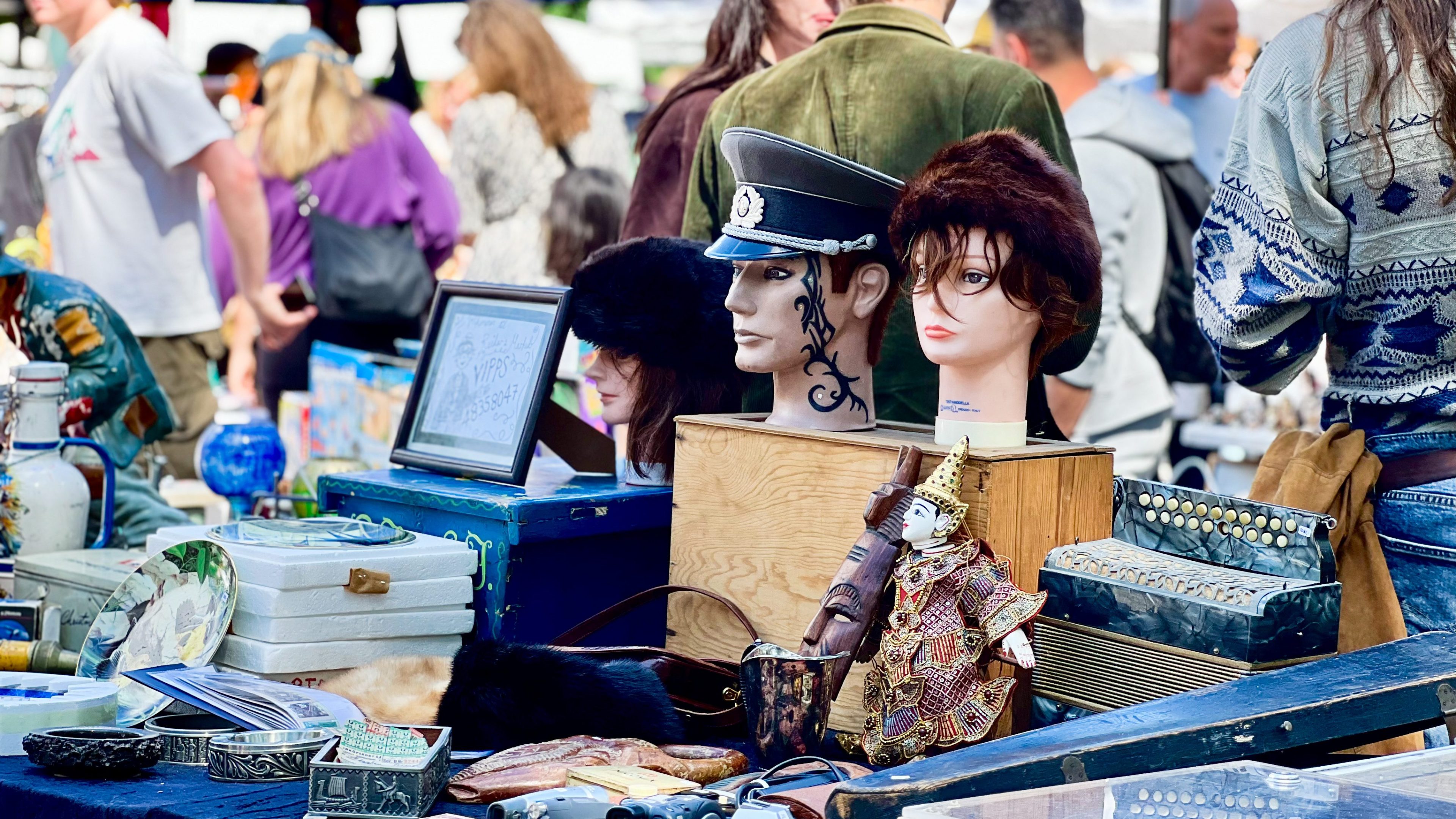 En loppemarkedsbod full av skattejakt viser utduker utsmykket med hatter, omgitt av vintagegjenstander og pyntegjenstander. I bakgrunnen ser shoppere på ulike gjenstander, hver på sin skattejakt.