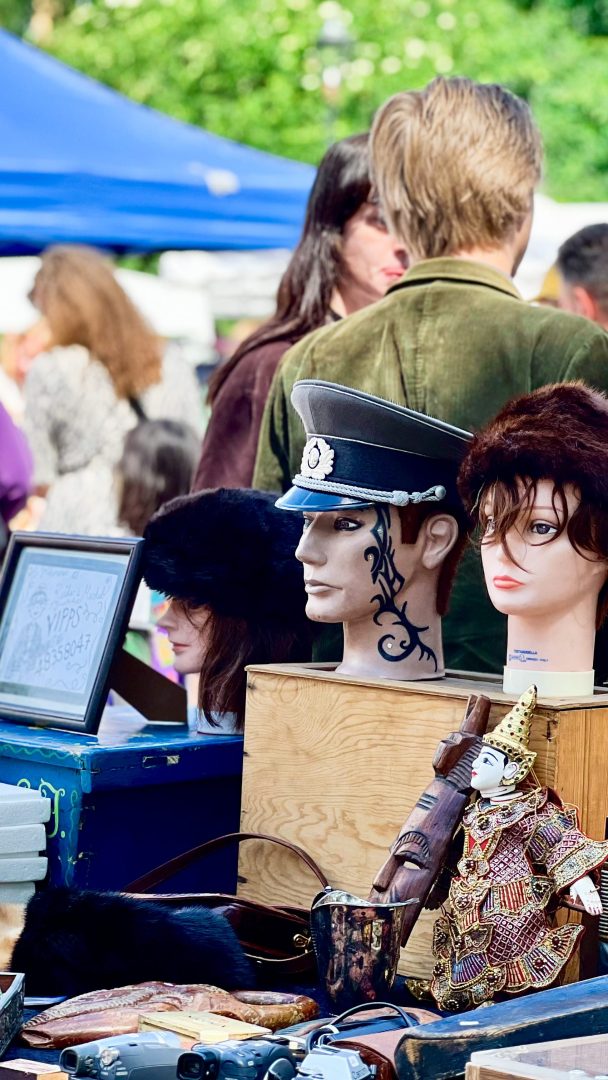 Utstillingsdukkehoder prydet med hatter vises på et bord på et utendørsmarked, der skattejaktentusiaster vandrer i bakgrunnen og leter etter skjulte skatter.