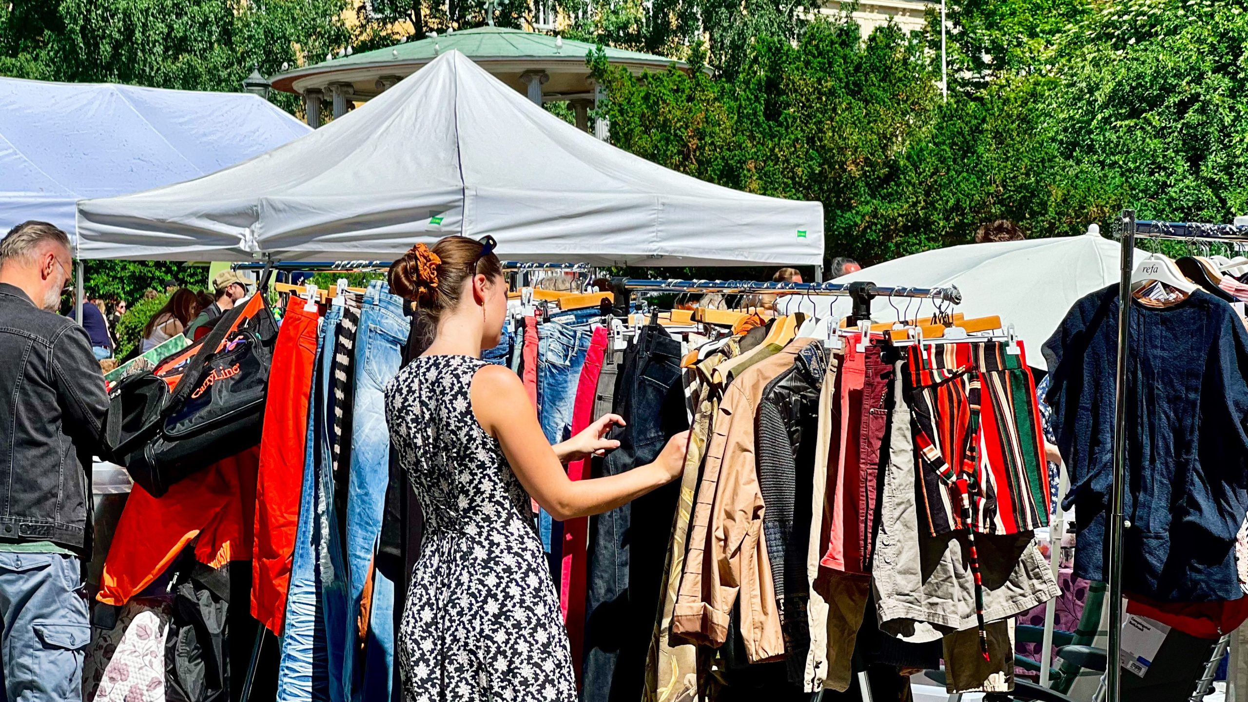 En kvinne legger ut på en skattejakt og surfer på klesstativer på et utendørsmarked med telt over hodet. Uformelle og formell klær i livlige farger vises, mens omkringliggende mennesker og grøntområder skaper et livlig bakteppe.