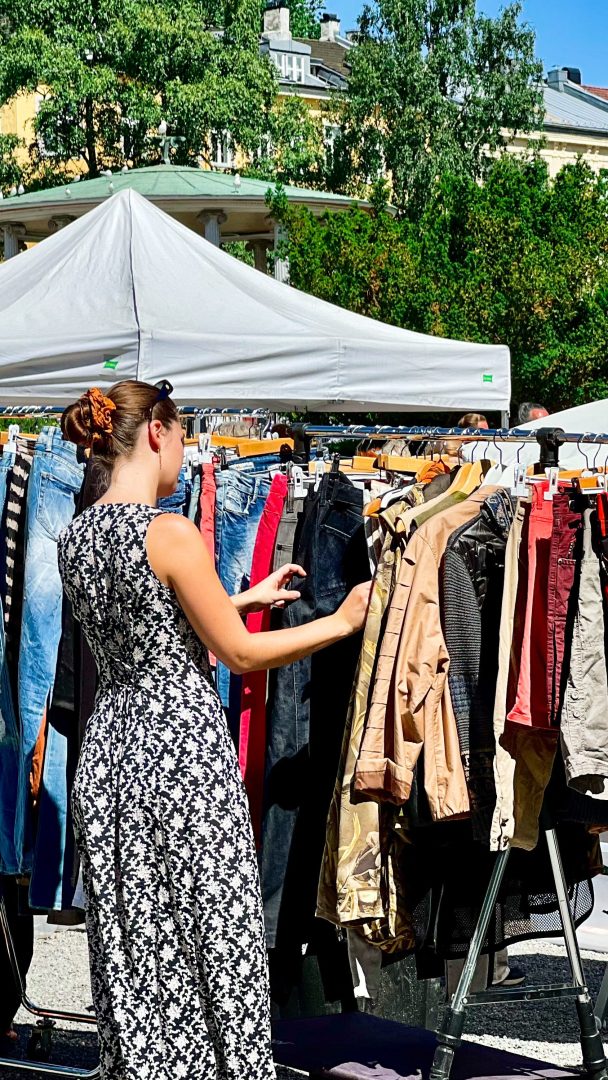 En kvinne i en blomsterkjole legger ut på en skattejakt og surfer på klesstativer på et utendørsmarked, med et hvitt telt og trær som gir det perfekte bakteppet.