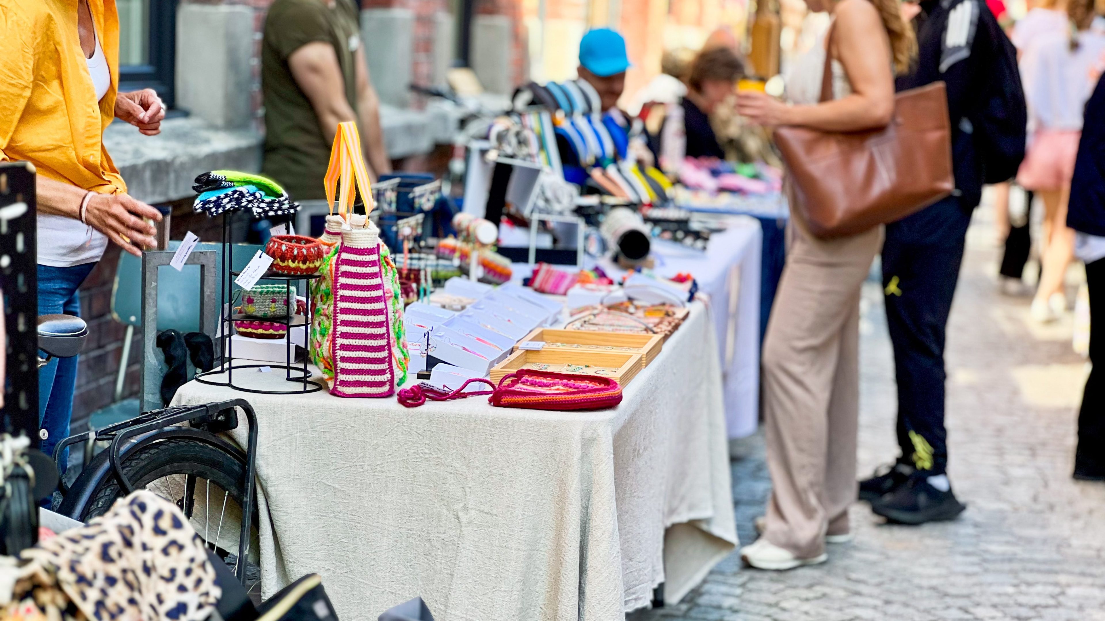 Utendørs markedsbod med en rekke håndlagde håndverk og tilbehør utstilt på et bord. Shoppere deltar i en skattejakt, og utforsker unike skatter mens folk surfer og handler i bakgrunnen.