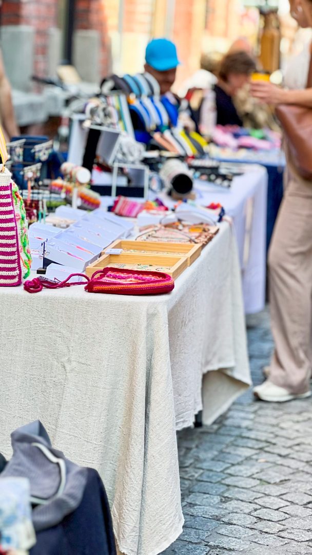 Utendørsmarkedsboden, som minner om en skattejakt, viser en rekke håndlagde armbånd og håndverk på en hvit duk. Besøkende blar ivrig gjennom gjenstandene, og hver oppdagelse øker spenningen ved jakten.