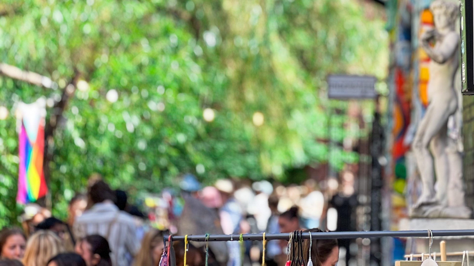 En livlig utendørs markedsscene utspiller seg mens folk engasjerer seg i en livlig skattejakt og leter gjennom boder. Et regnbueflagg og diverse hengende gjenstander fanger blikket, mens en statue står stolt i bakgrunnen. Lyse grøntområder omslutter området, og bidrar til det fargerike veggteppet på dette travle stedet.