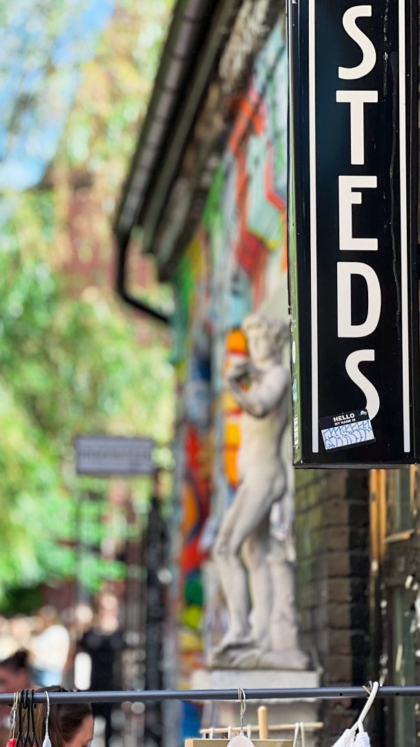 Uteområdet er markert med et svart skilt som delvis viser «STEDS», og legger til rette for et skattejakteventyr. I den uskarpe bakgrunnen gir et fargerikt veggmaleri og en klassisk statue et kunstnerisk preg til dette spennende stedet.