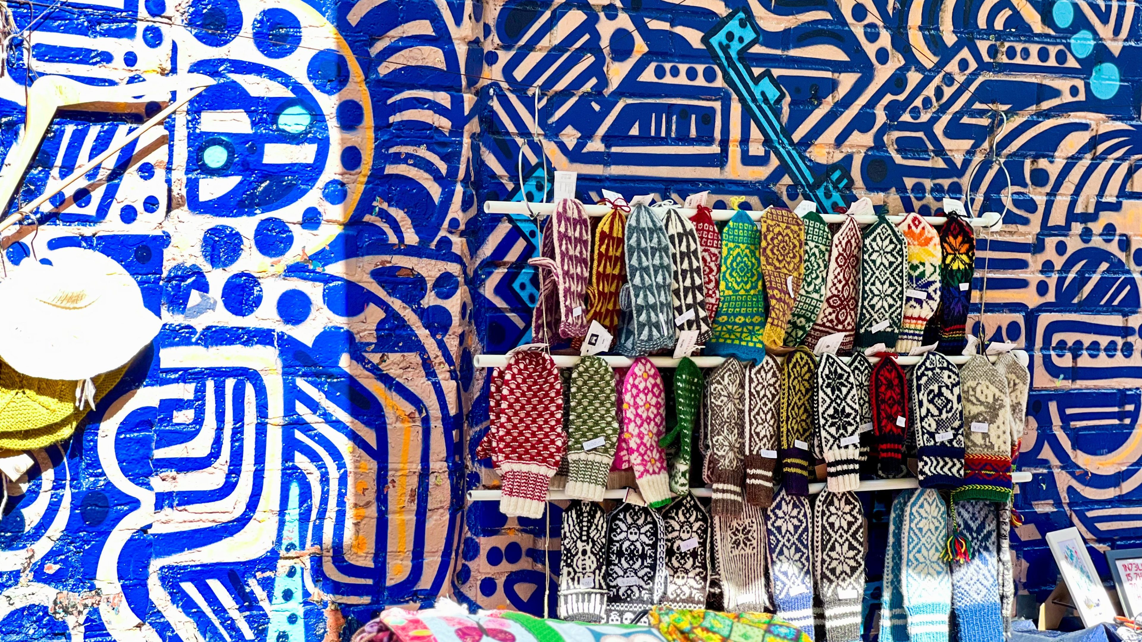 Fargerike strikkevotter på stativ inviterer til skattejakt foran et levende blått og oransje geometrisk veggmaleri. Hatter henger fristende på venstre side, og bidrar til skattejakten.