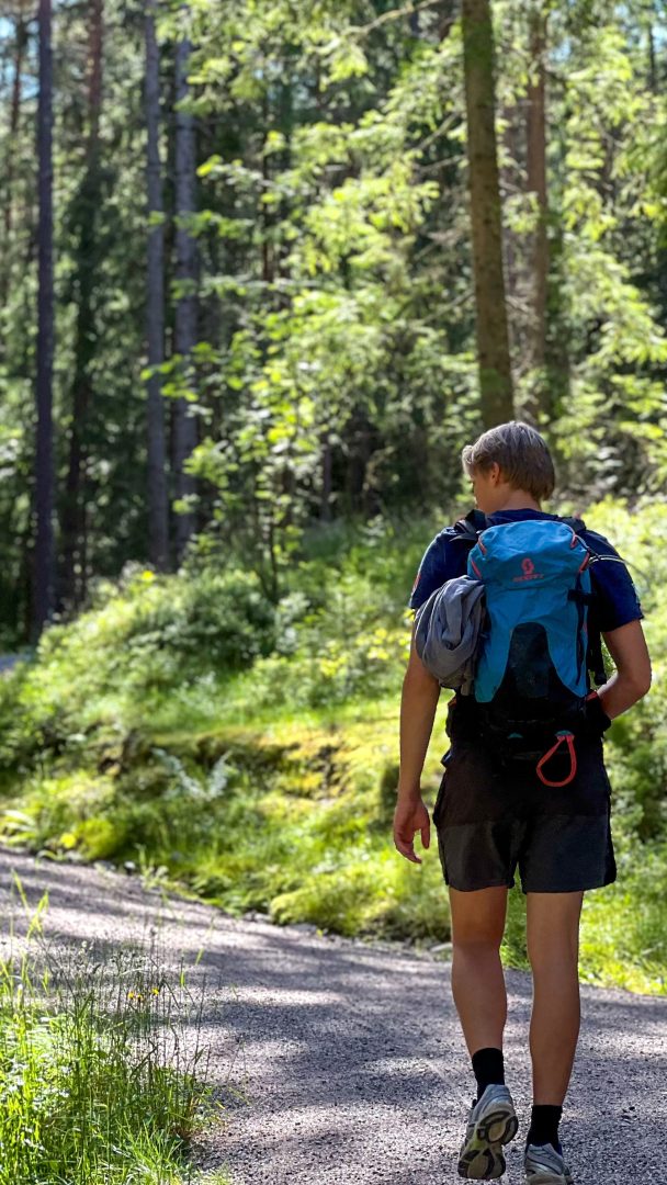 En person med en blå ryggsekk legger ut på et topptur-eventyr langs en skogssti, omgitt av høye trær og flekkete sollys som filtrerer gjennom bladene.