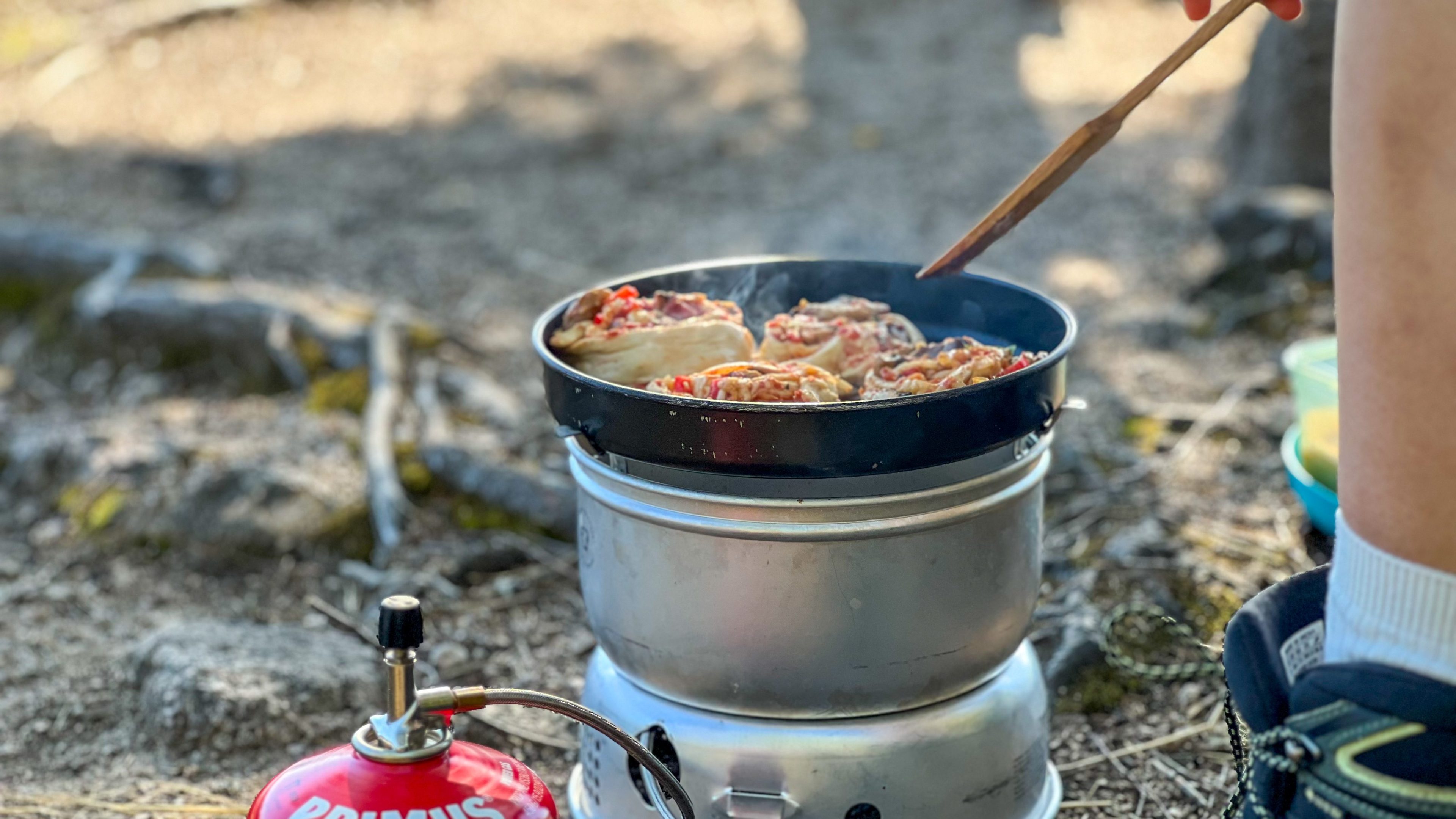 En person lager dyktig bakverk i en panne på en bærbar campingkomfyr under et naturskjønn topptur-eventyr. En gassbeholder hviler i nærheten, og fullfører det perfekte utendørs kulinariske oppsettet.