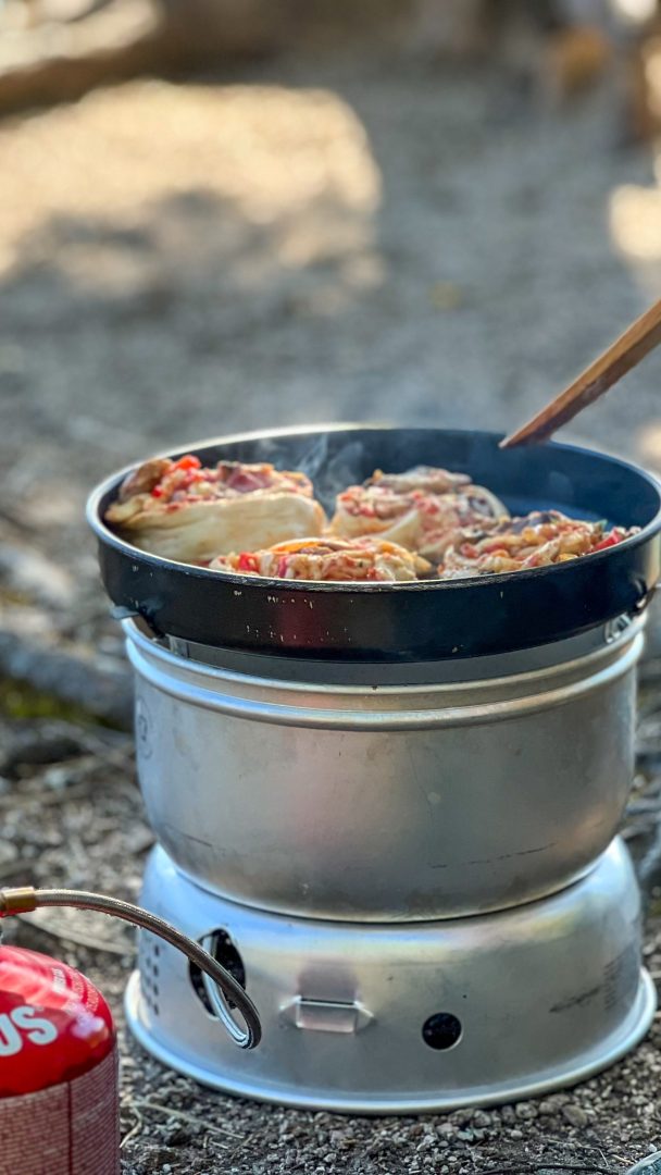 Kanelsnurrer syder i en stekepanne på en bærbar gasskomfyr under et topptur-eventyr, med en rød gassbeholder like ved.