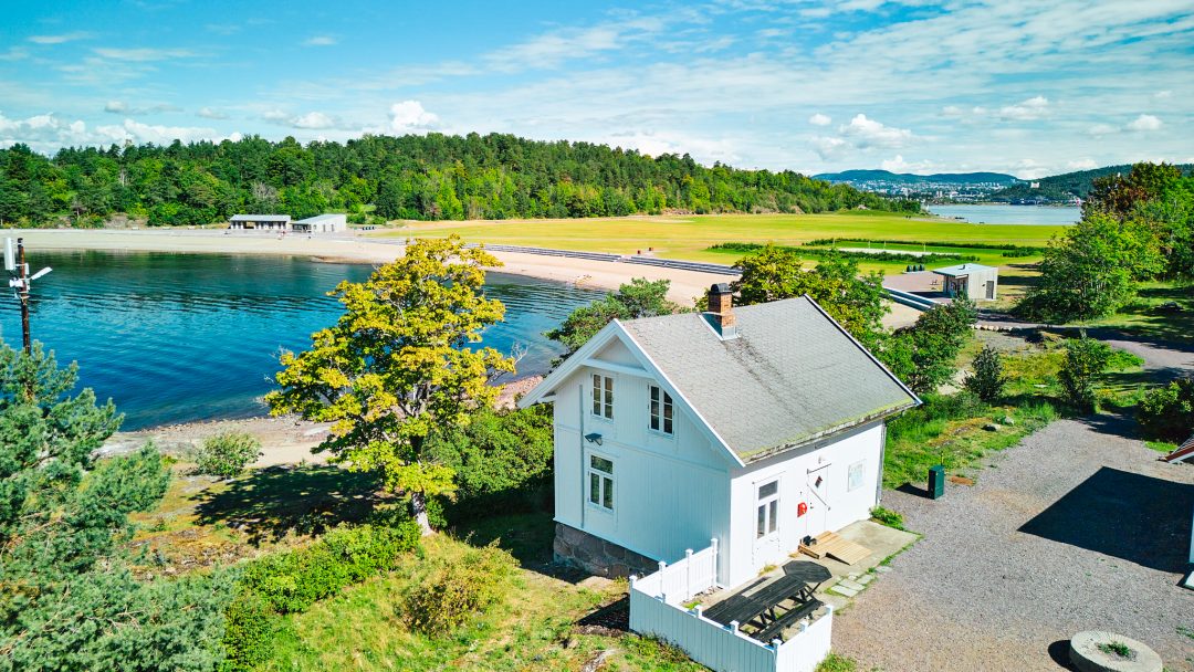Et sjarmerende hvitt hus ved en strand og frodige grøntområder under en klarblå himmel, med fjerne åser mot bakgrunn av glitrende vann.