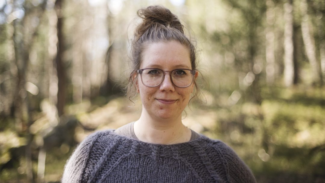 En person med briller og bollefrisyre, kledd i en grå genser, står utendørs blant slørete trær og tenker kanskje på klimaregnskapet sitt.