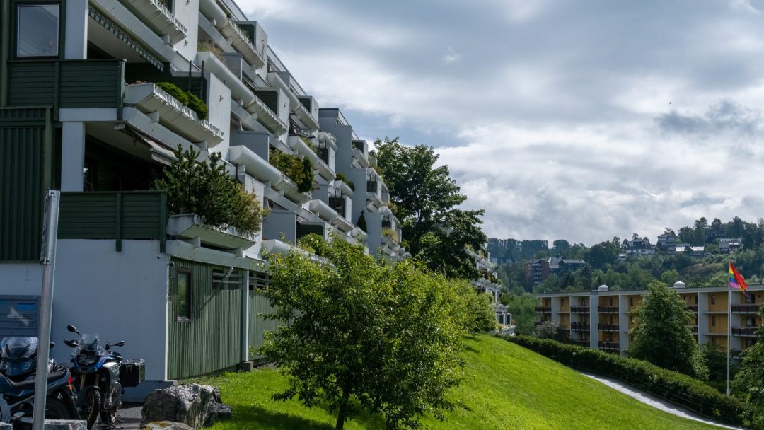 En rekke moderne leilighetsbygg i flere etasjer med balkonger og solcellepaneler, omgitt av grøntområder under en delvis overskyet himmel. Et flagg er synlig i bakgrunnen.
