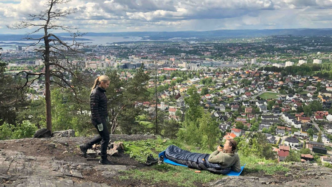 To personer på en steinete høyde med utsikt over et bybilde, den ene står i friluftsutstyr ved siden av telten, mens den andre hviler på en blå campingmatte i en sovepose. En overskyet himmel ruver over.