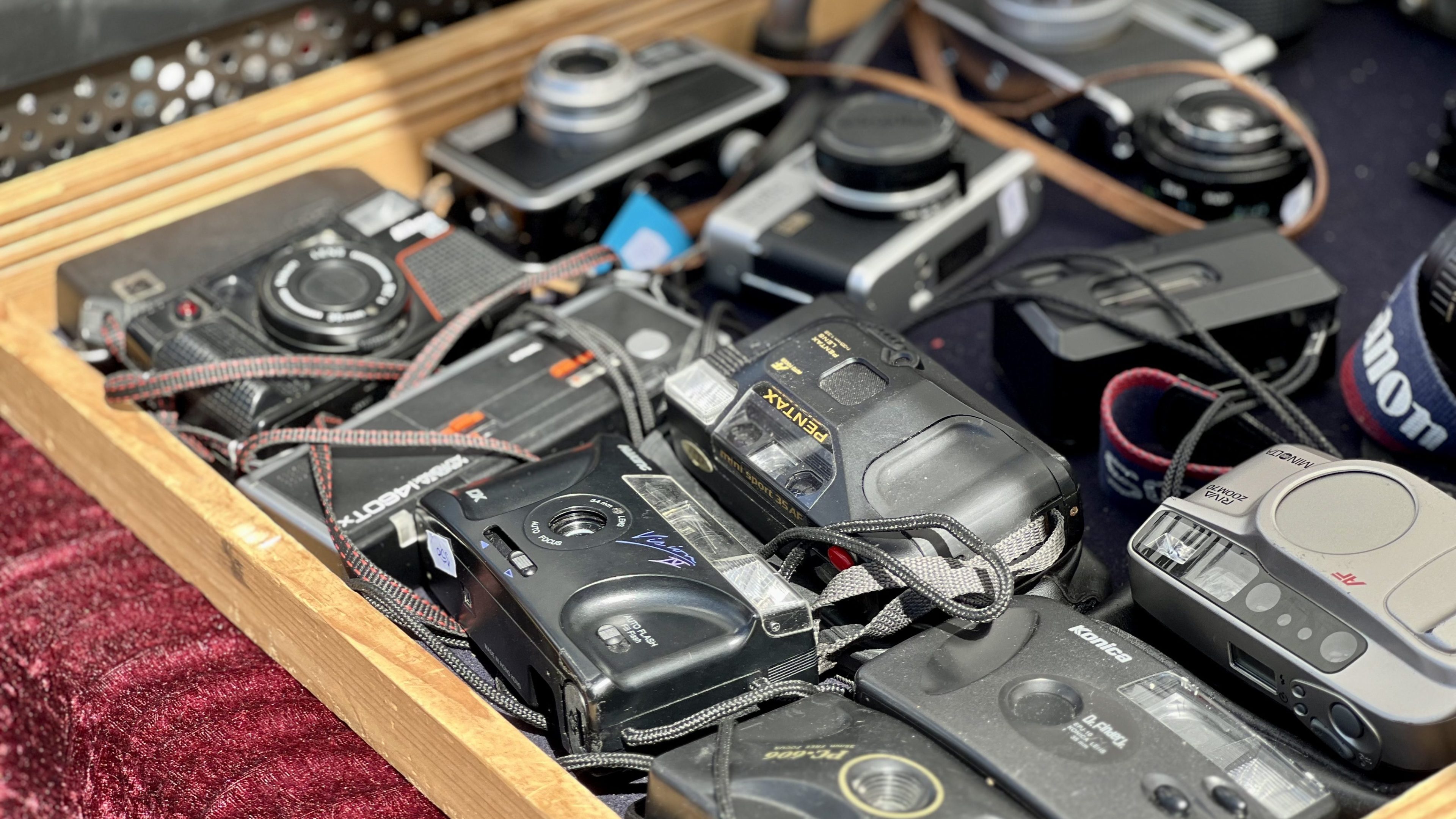 En treboks, som en skattejakt, er fylt med forskjellige vintage filmkameraer, inkludert merker som Canon og Revue, med stropper festet, alle sitter på et burgunder stoff.