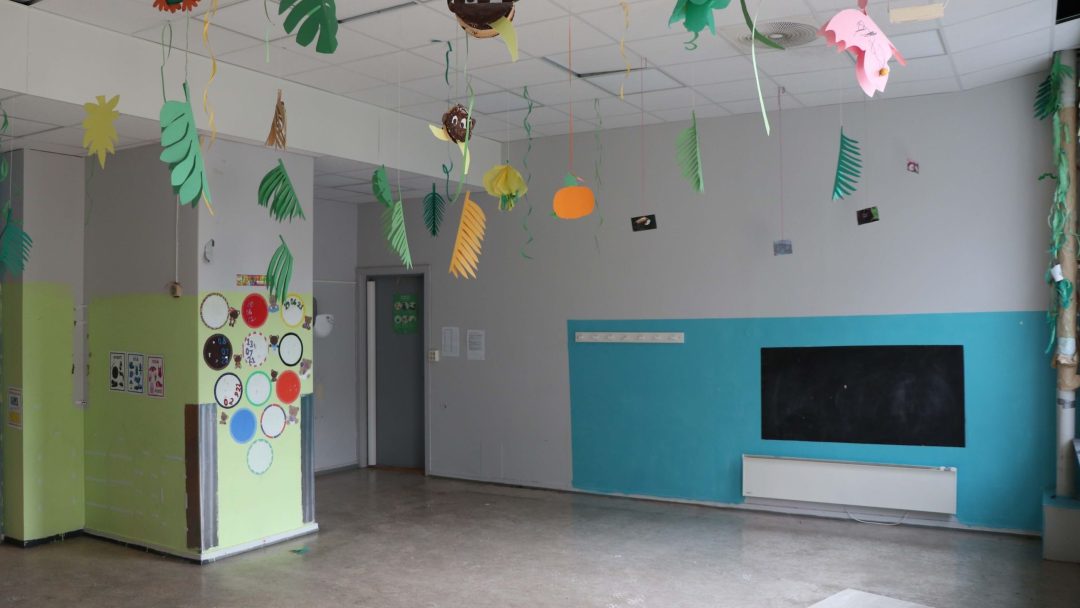 Et tomt klasserom, utsmykket med fargerike papirdekorasjoner som dingler fra taket, har en tavle på veggen. På venstre side viser en sirkulær skjerm forskjellige fargede sirkler, som subtilt minner oss om indirekte utslipp og deres innvirkning på vårt daglige miljø.