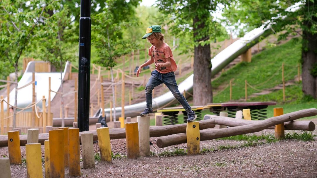 På rolige lekeplasser går et barn langs trebjelker innrammet av frodige trær og grøntområder, med sklier synlige i bakgrunnen.