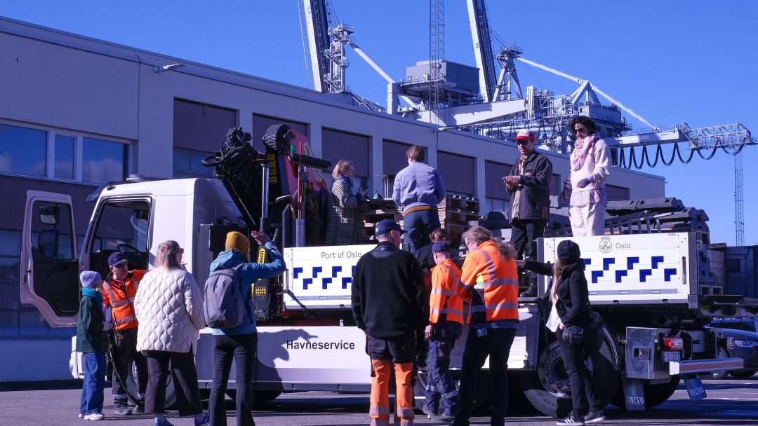 En gruppe mennesker, noen i sikkerhetsvest, står med stolthet på og rundt en lastebil merket «Havneservice» ved en havn, med kraner i bakgrunnen.