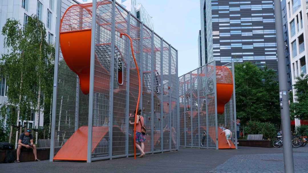 En person lener seg mot en høy, oransje, burlignende offentlig kunststruktur som ligner pulserende lekeplasser i urbane omgivelser, omgitt av moderne bygninger og trær.