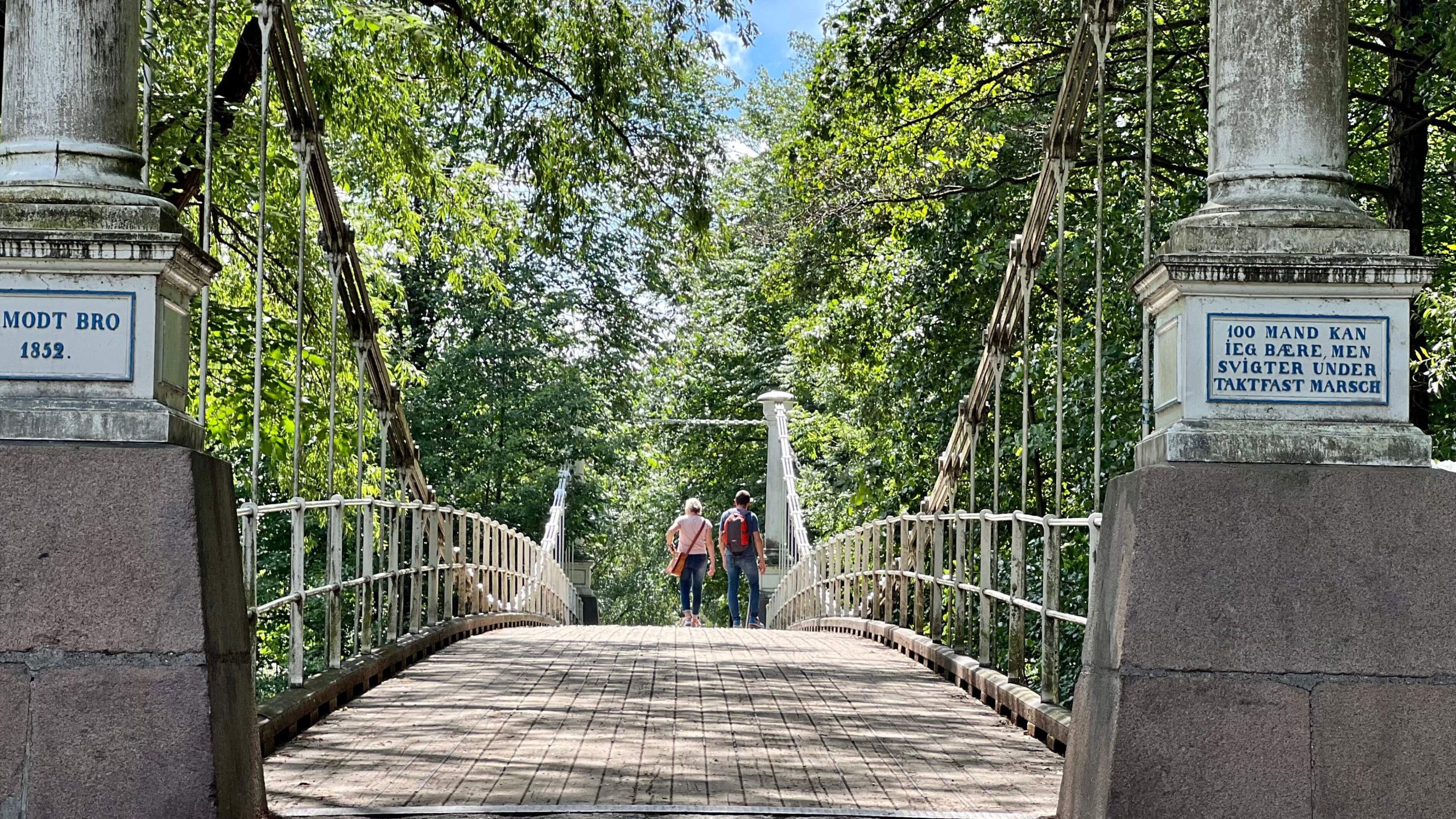 To personer som rusler på en hengebro i tre, omfavnet av frodige trær. I nærheten avslører skilt på steinsøylene tekst på et annet språk, som antyder skjulte sykkelstier som venter på å bli oppdaget.