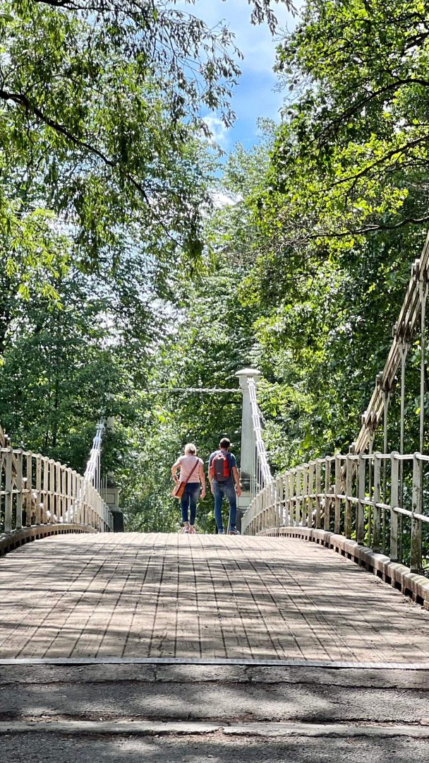 To personer som går på en metallbro omgitt av frodige grønne trær under en delvis overskyet himmel, hvor sykkelstier vever seg grasiøst gjennom landskapet.